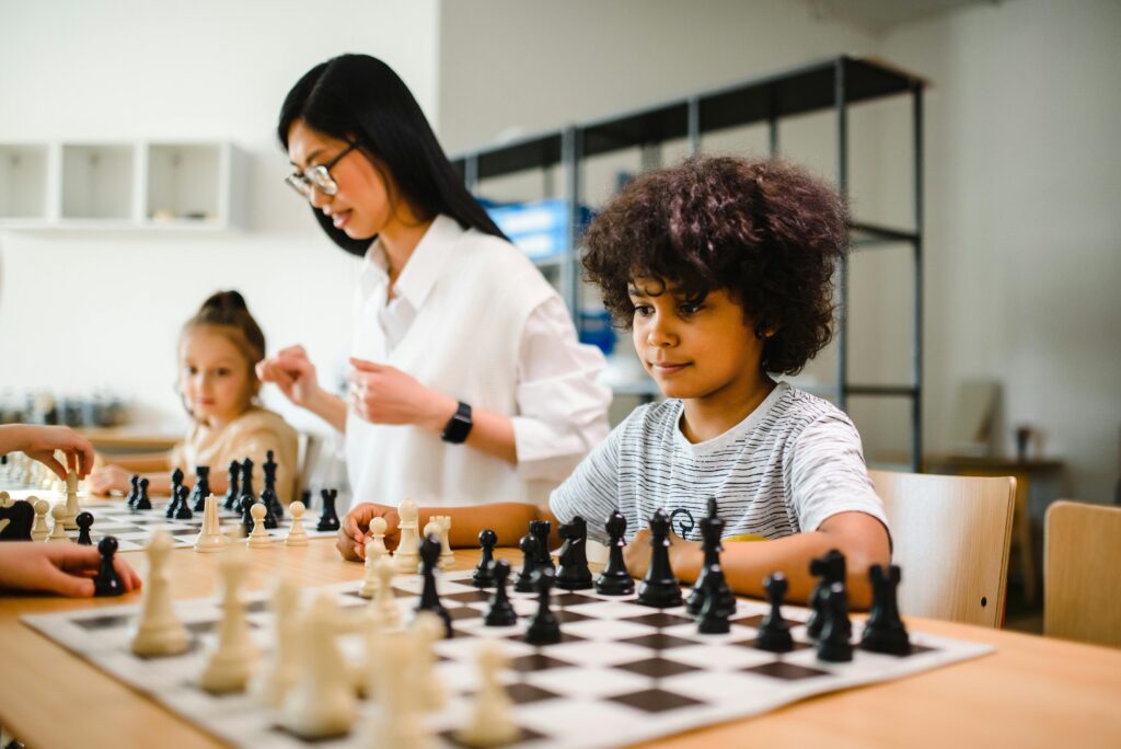A multicultural group of children playing chess under teacher's guidance in a classroom setting.